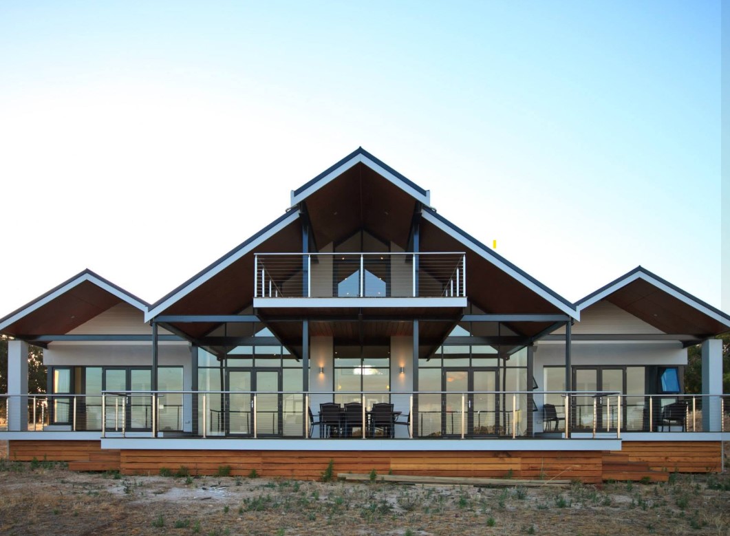 Rural pavilion home at dusk with three gabled roofs, timber-lined eaves and a full-width deck