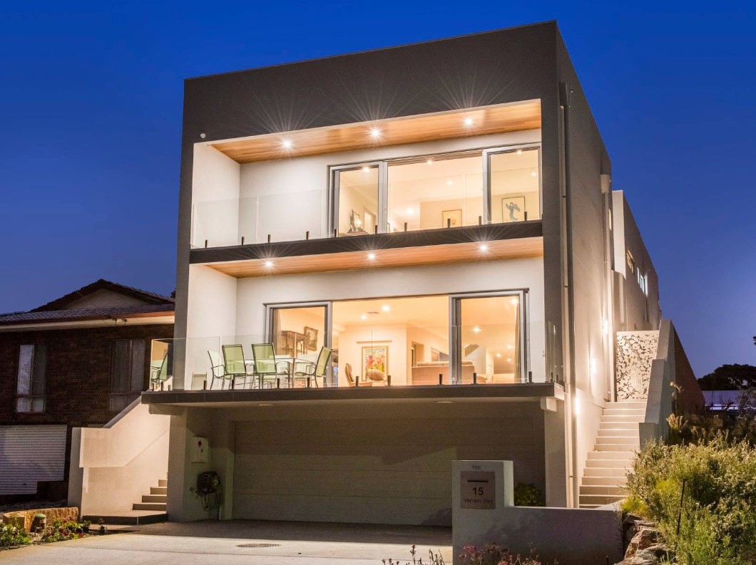 Custom two-storey eco home lit at dusk, with timber-lined balcony soffits and glass balustrades