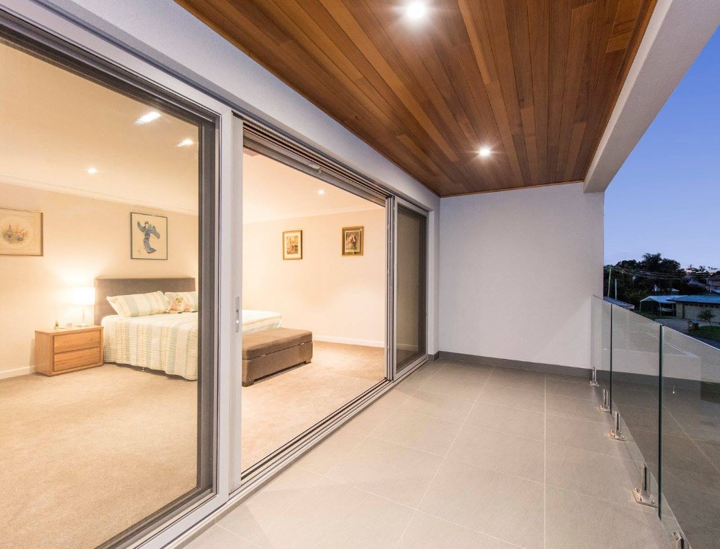 Master bedroom opening onto a tiled balcony with a timber-lined ceiling and glass balustrade